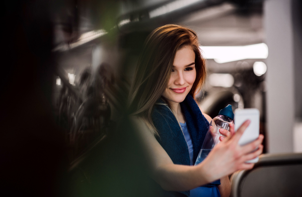 A portrait of young girl or woman with water bottle and smartphone in a gym, taking selfie.