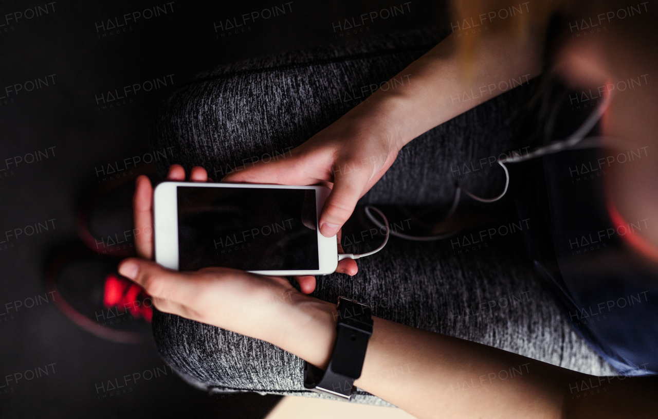 A midsection of young girl or woman with earphones and smartphone in a gym, a top view.