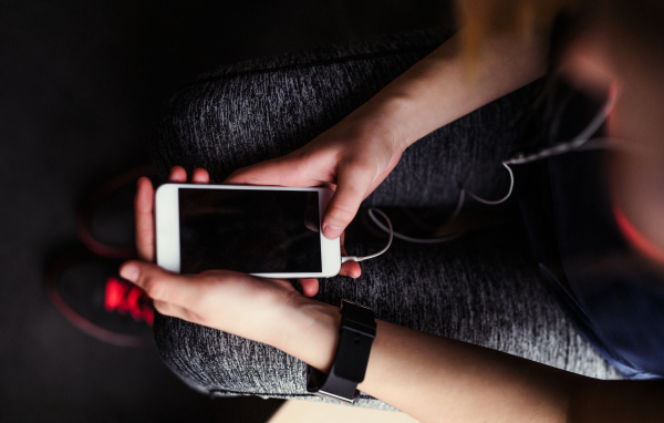 A midsection of young girl or woman with earphones and smartphone in a gym, a top view.