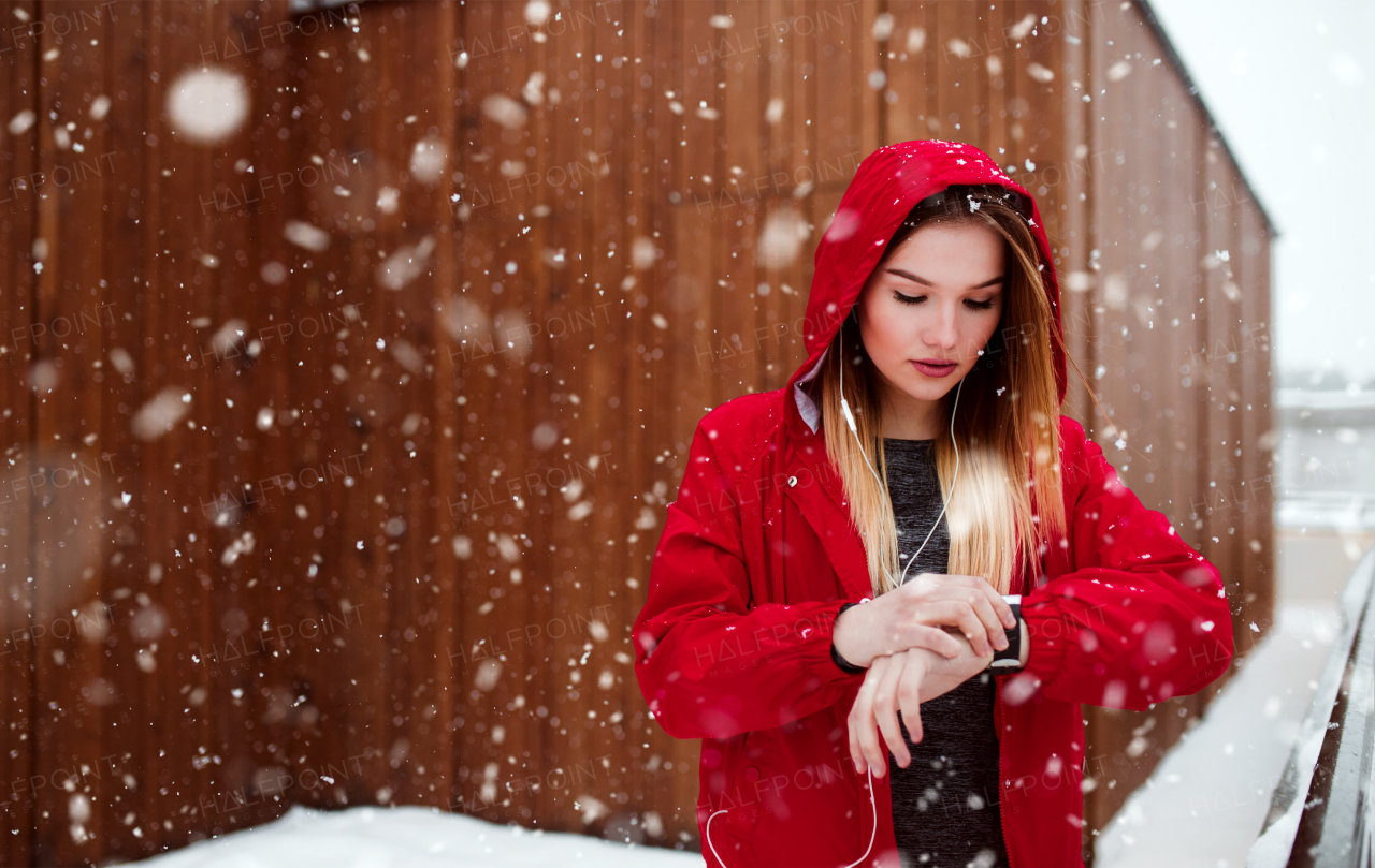 A portrait of young girl or woman with earphones using smartwatch outdoors in winter.