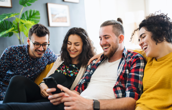 A group of young cheerful friends with smartphone sitting on sofa indoors, house sharing concept.