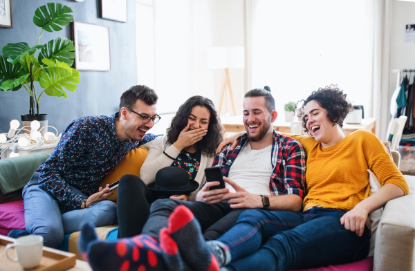 A group of young cheerful friends with smartphone sitting on sofa indoors, house sharing concept.