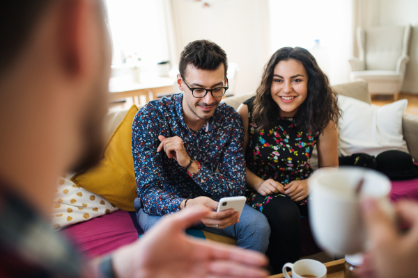 A group of young cheerful friends with smartphone sitting on sofa indoors, house sharing concept.