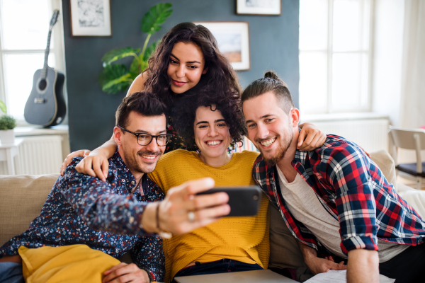 A group of young cheerful friends with smartphone sitting on sofa indoors, taking selfie. House sharing concept.