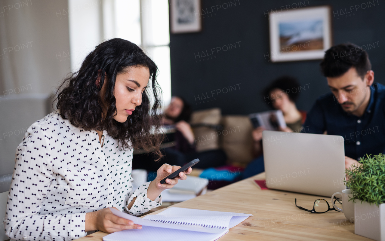 A group of young cheerful friends with smartphone and laptop sitting at table indoors, house sharing concept.