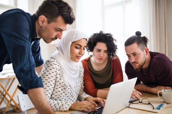 A multi-ethnic group of young cheerful friends with laptop sitting at table indoors, house sharing concept.