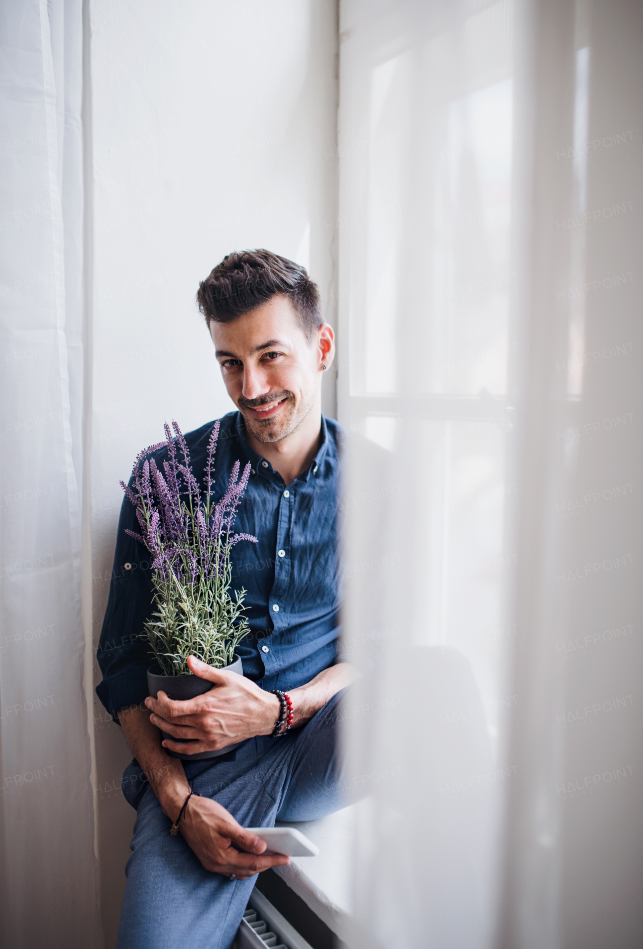 A portrait of young man sitting on window sill indoors, holding a plant. Copy space.