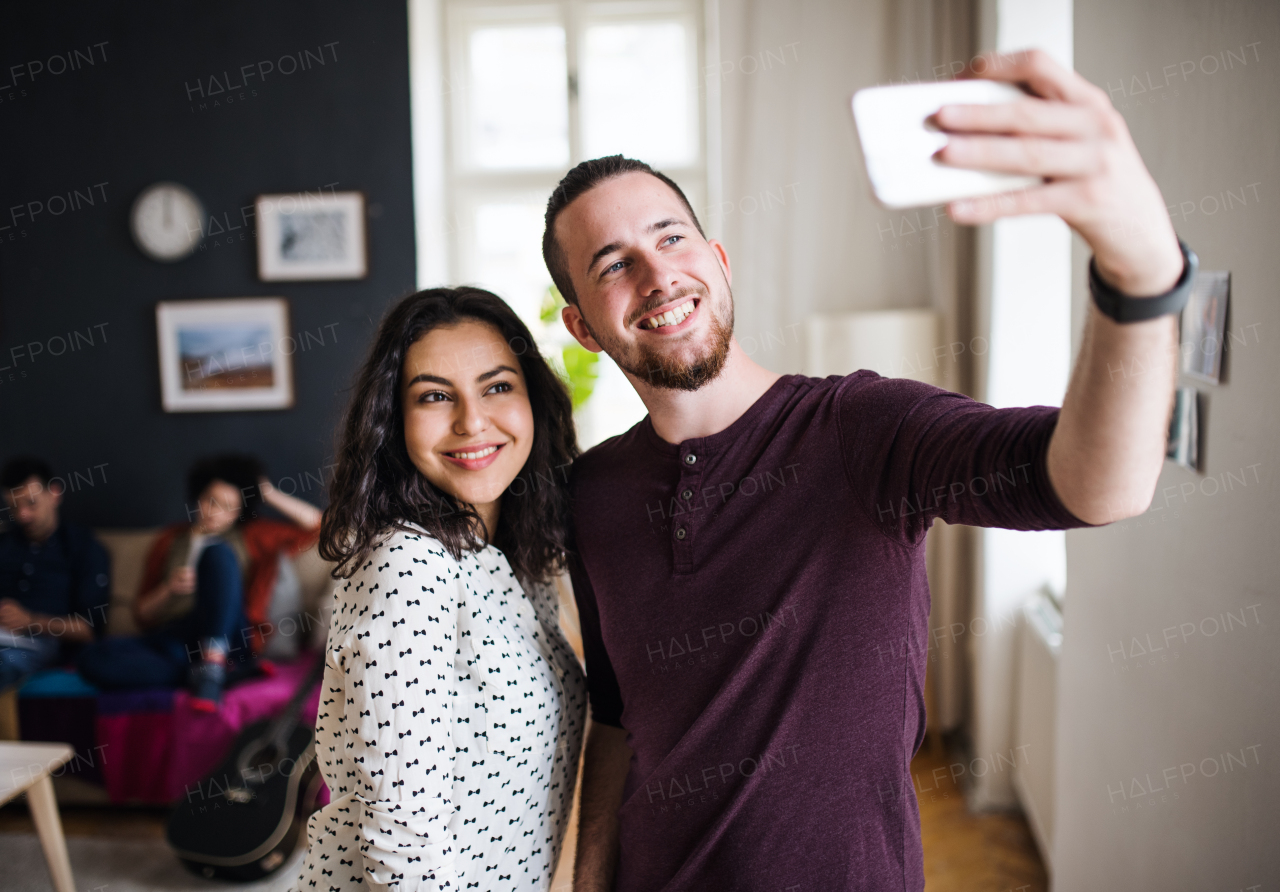 A group of young cheerful friends with smartphone indoors, taking selfie. House sharing concept.