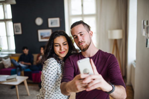 A group of young cheerful friends with smartphone indoors, taking selfie. House sharing concept.