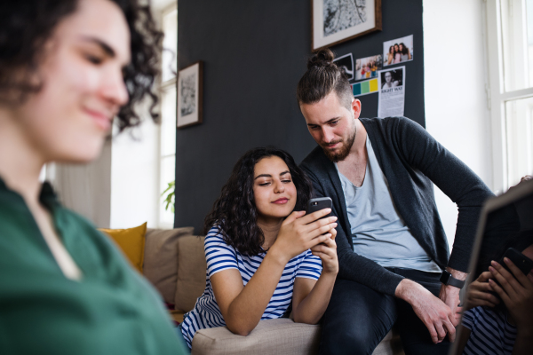 A group of young cheerful friends with smartphone sitting on sofa indoors, house sharing concept.