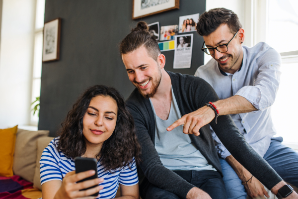 A group of young cheerful friends with smartphone sitting on sofa indoors, house sharing concept.