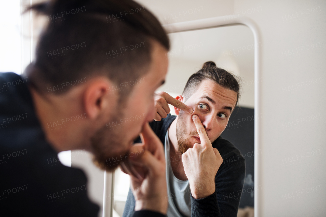 A young hipster man indoors looking in the mirror, squeezing a pimple. Acne problem.