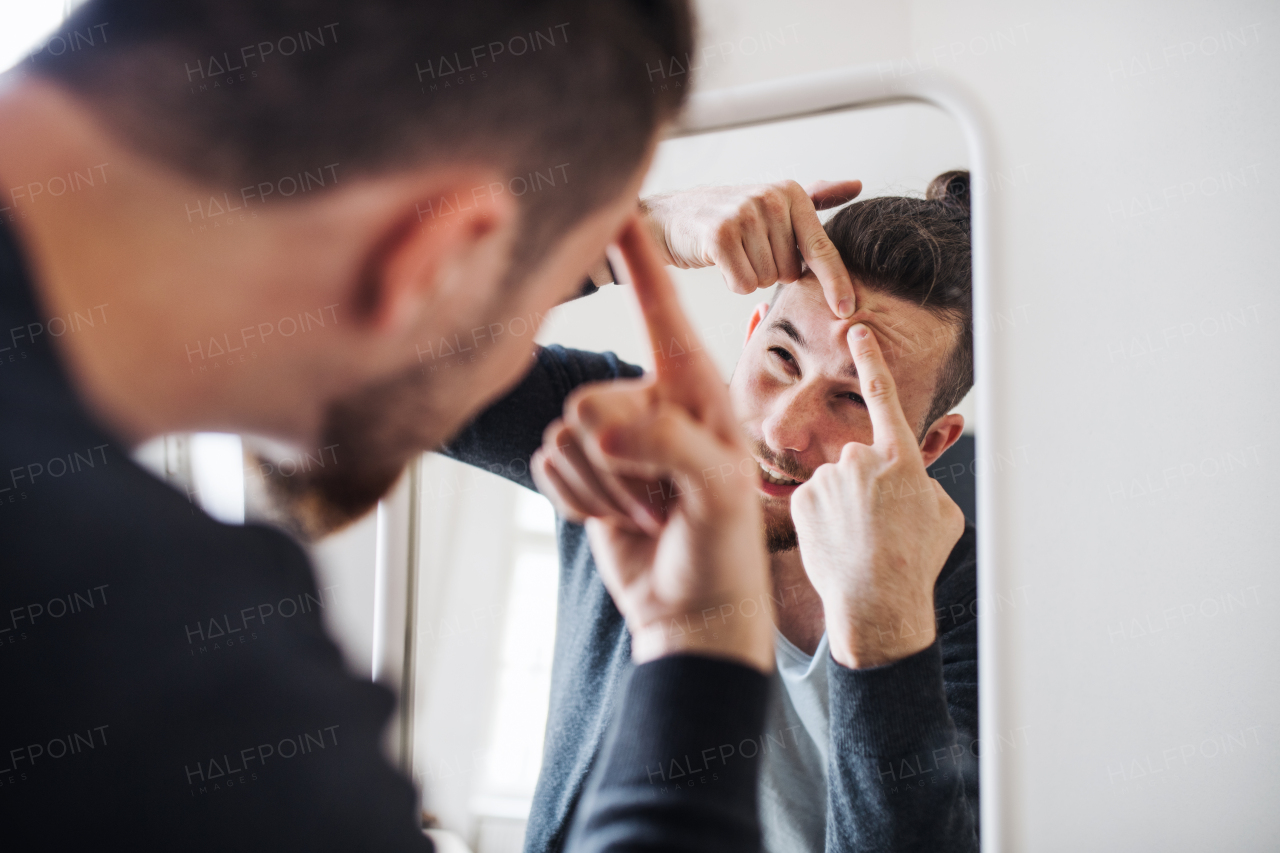 A young hipster man indoors looking in the mirror, squeezing a pimple. Acne problem.