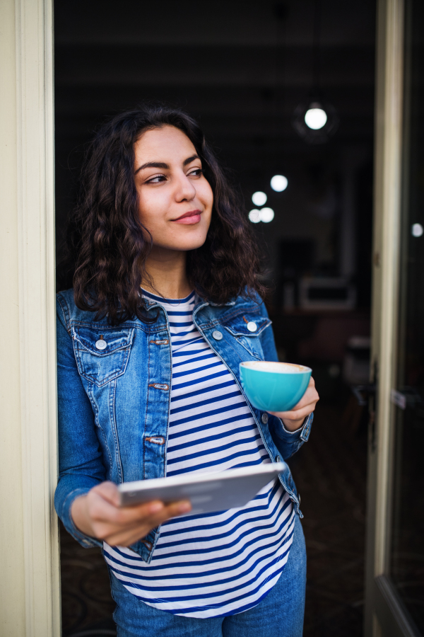 A beautiful young woman with tablet and coffee standing at the front door in cafe, waiting for somebody.