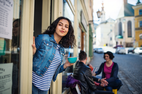 A beautiful young woman with coffee standing at the front door in cafe, looking for somebody.