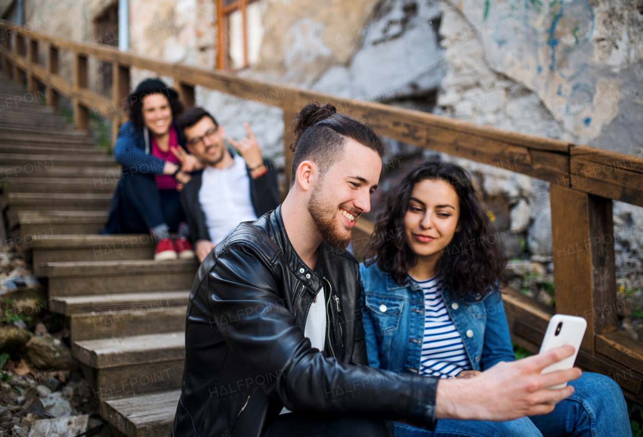 A group of young friends with smartphone sitting outdoors on staircase in town, taking selfie.