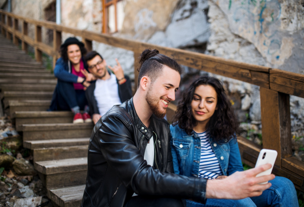 A group of young friends with smartphone sitting outdoors on staircase in town, taking selfie.