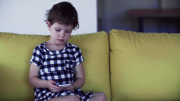 A cute small girl sitting on sofa indoors at home, playing with smartphone. Slow motion.