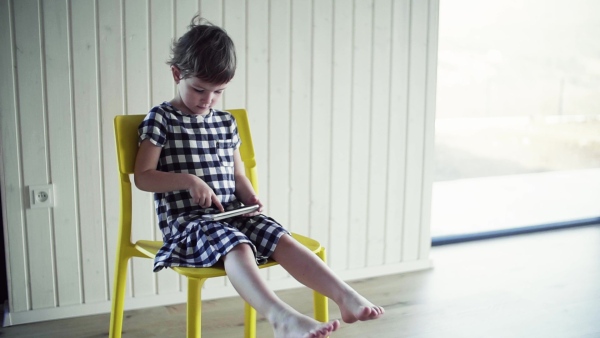 A cute small girl sitting on chair indoors at home, playing with smartphone. Slow motion.