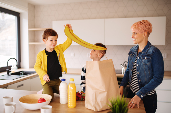 A happy young woman with two children unpacking shopping in a kitchen.