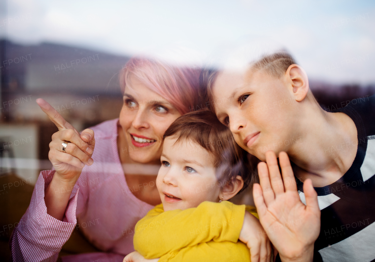 A portrait of young woman with two children at home, looking out of window. Shot through glass.