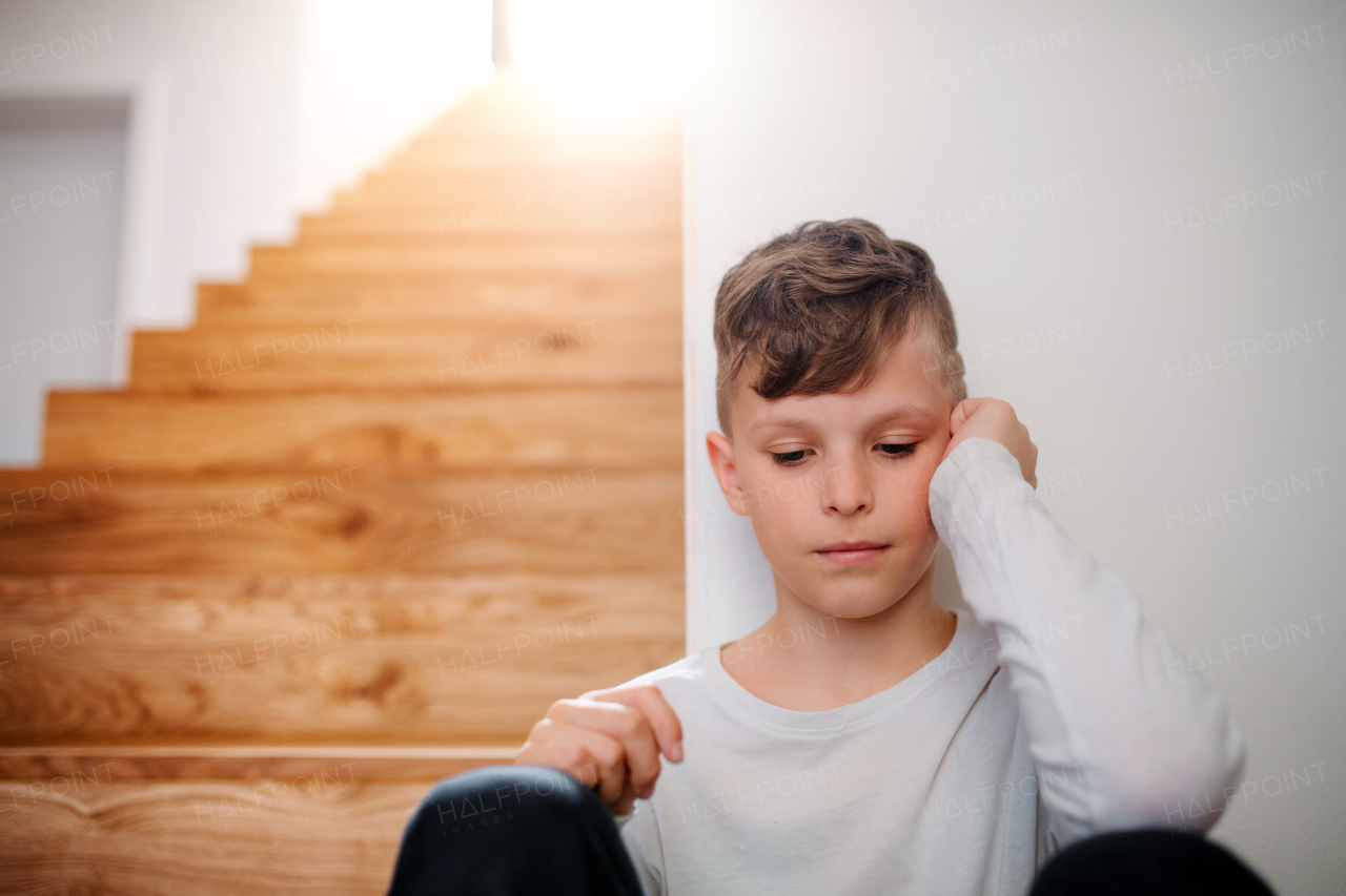 A sad small unhappy boy sitting on the floor by the stairs. Copy space.