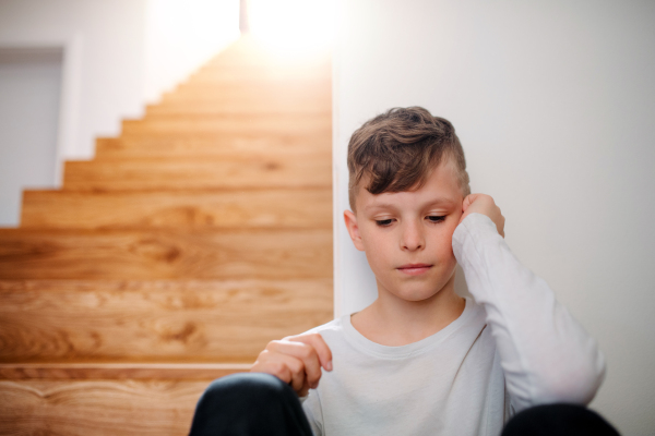 A sad small unhappy boy sitting on the floor by the stairs. Copy space.