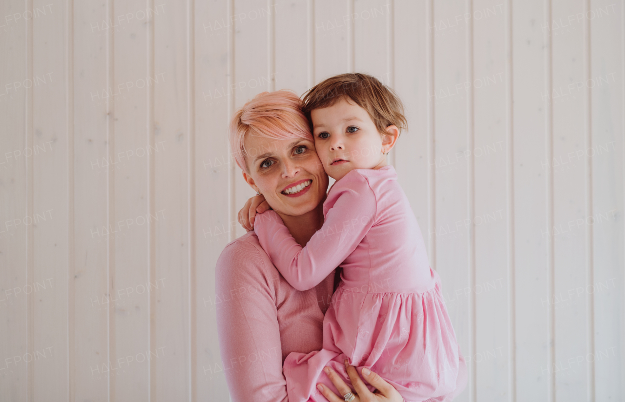 A portrait of young woman holding small daughter, white wooden wall in the background.