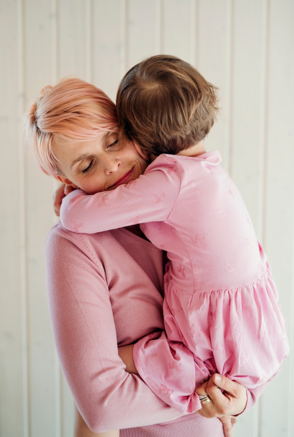 A portrait of young woman holding small daughter, white wooden wall in the background.