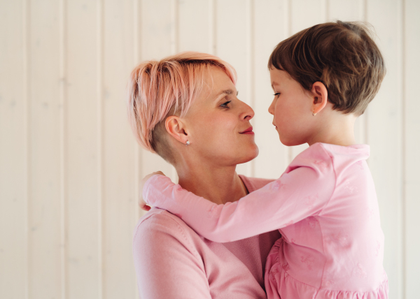 A portrait of young woman holding small daughter, white wooden wall in the background.