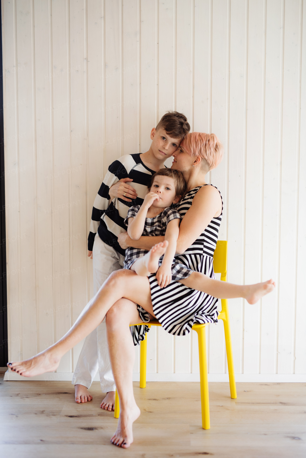 Young woman with two children in black and white clothes sitting on yellow chair against white wooden wall.