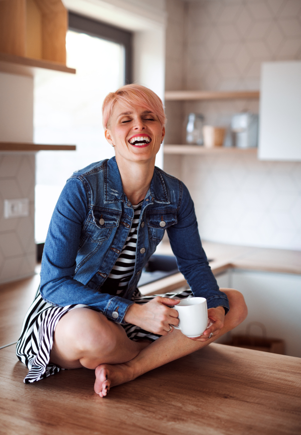 A young attractive woman with a coffee sitting on a counter in a kitchen at home, laughing.