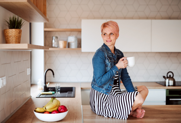 A young attractive woman with a coffee sitting on a counter in a kitchen at home.