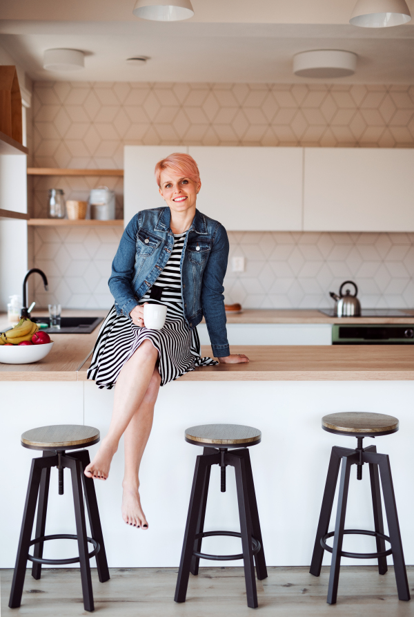 A young attractive woman with a coffee sitting on a counter in a kitchen at home.