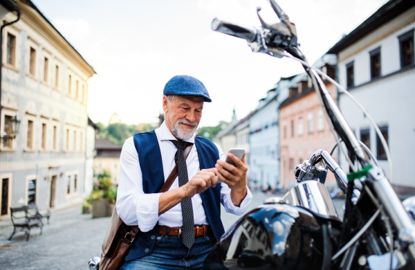 A cheerful senior businessman with motorbike in town, using smartphone.