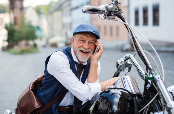 A cheerful senior businessman with motorbike in town, using smartphone.