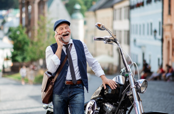 A cheerful senior businessman with motorbike in town, using smartphone.