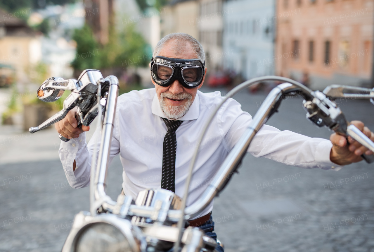 A front view of senior businessman with goggles and motorbike in town, looking at camera.