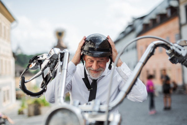 A happy senior businessman with motorbike in town, putting on helmet.