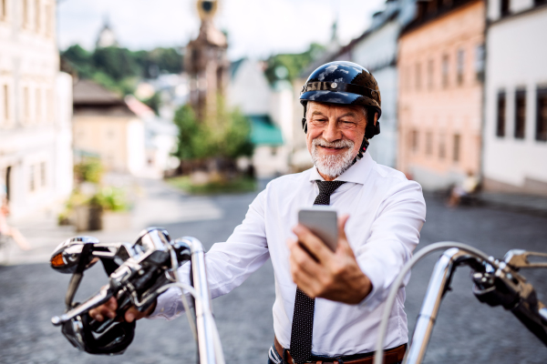 A cheerful senior businessman with motorbike in town, using smartphone.