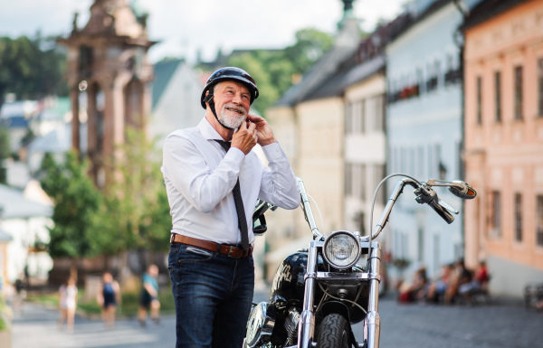 A cheeful senior businessman with motorbike in town, putting on helmet.