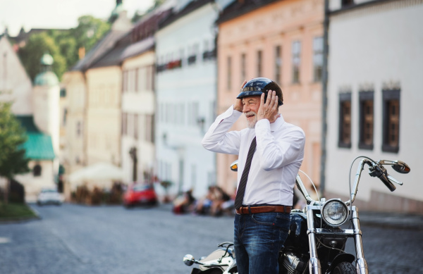 A cheeful senior businessman with motorbike in town, putting on helmet.