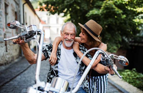 A front view of cheerful senior couple travellers with motorbike in town.