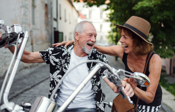 A front view of cheerful senior couple travellers with motorbike in town.