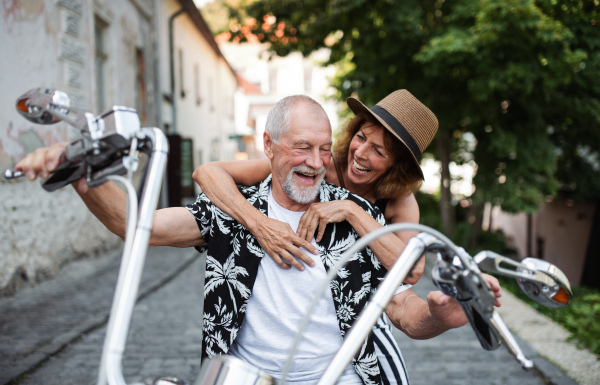 A front view of cheerful senior couple travellers with motorbike in town.