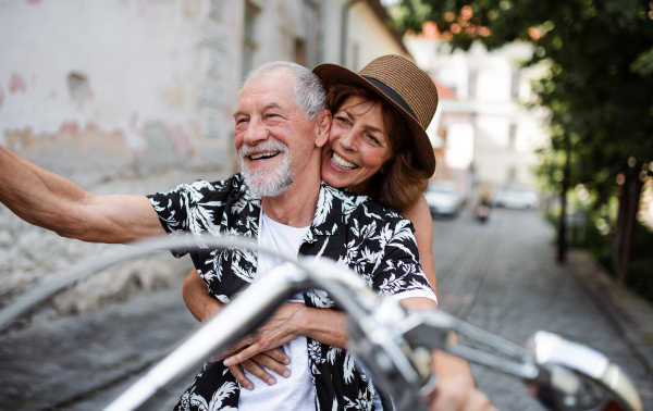 A front view of cheerful senior couple travellers with motorbike in town.