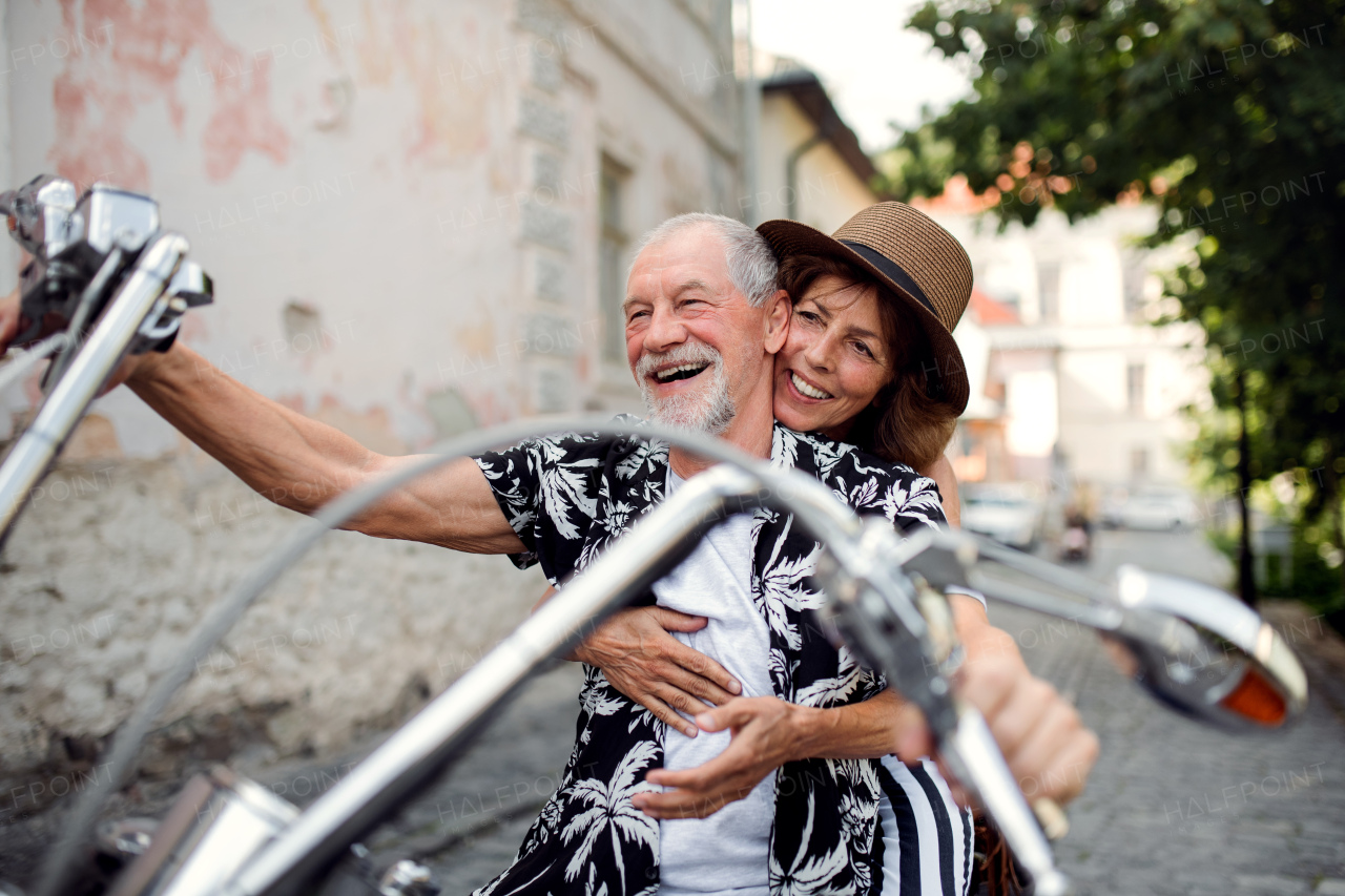 A front view of cheerful senior couple travellers with motorbike in town.