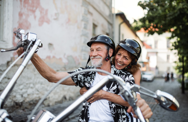 A front view of cheerful senior couple travellers with motorbike in town.