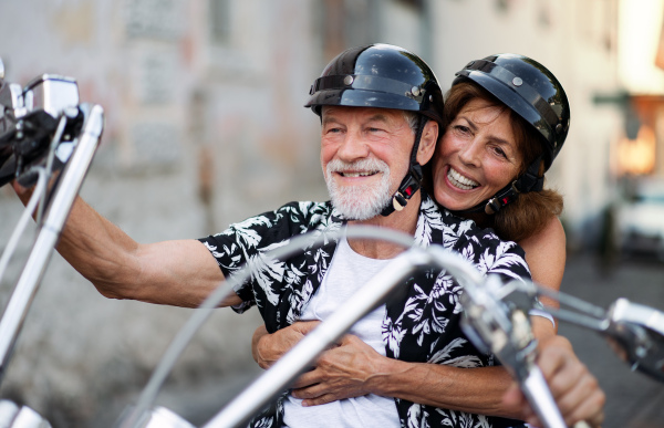 A front view of cheerful senior couple travellers with motorbike in town.