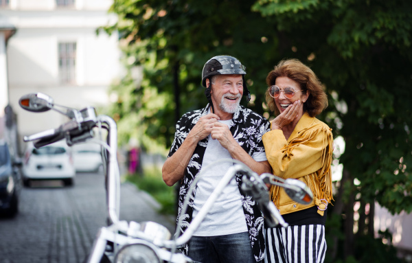 A front view of cheerful senior couple travellers with motorbike in town.